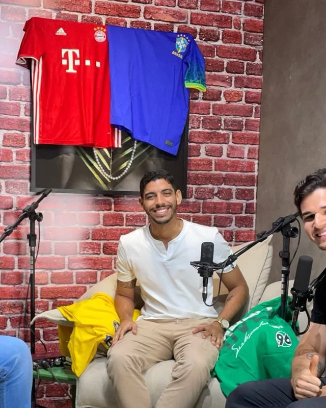 Victor Hugo Sales Santos sorrindo durante a gravação de um podcast sobre esporte, com camisas de futebol na parede